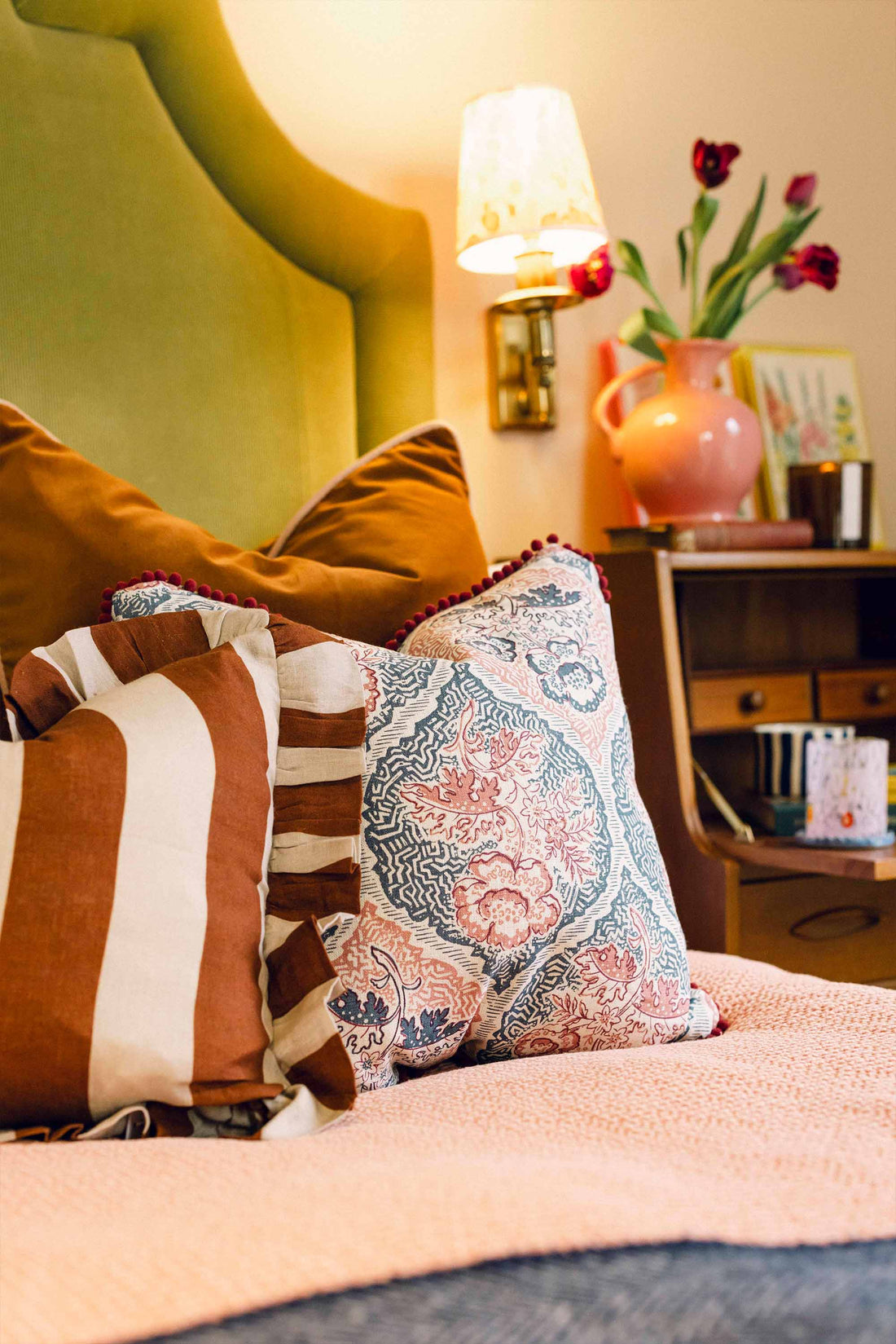 Decorative pillows on a bed with a lamp and vase of flowers in the background. LAMP LDN LIVING