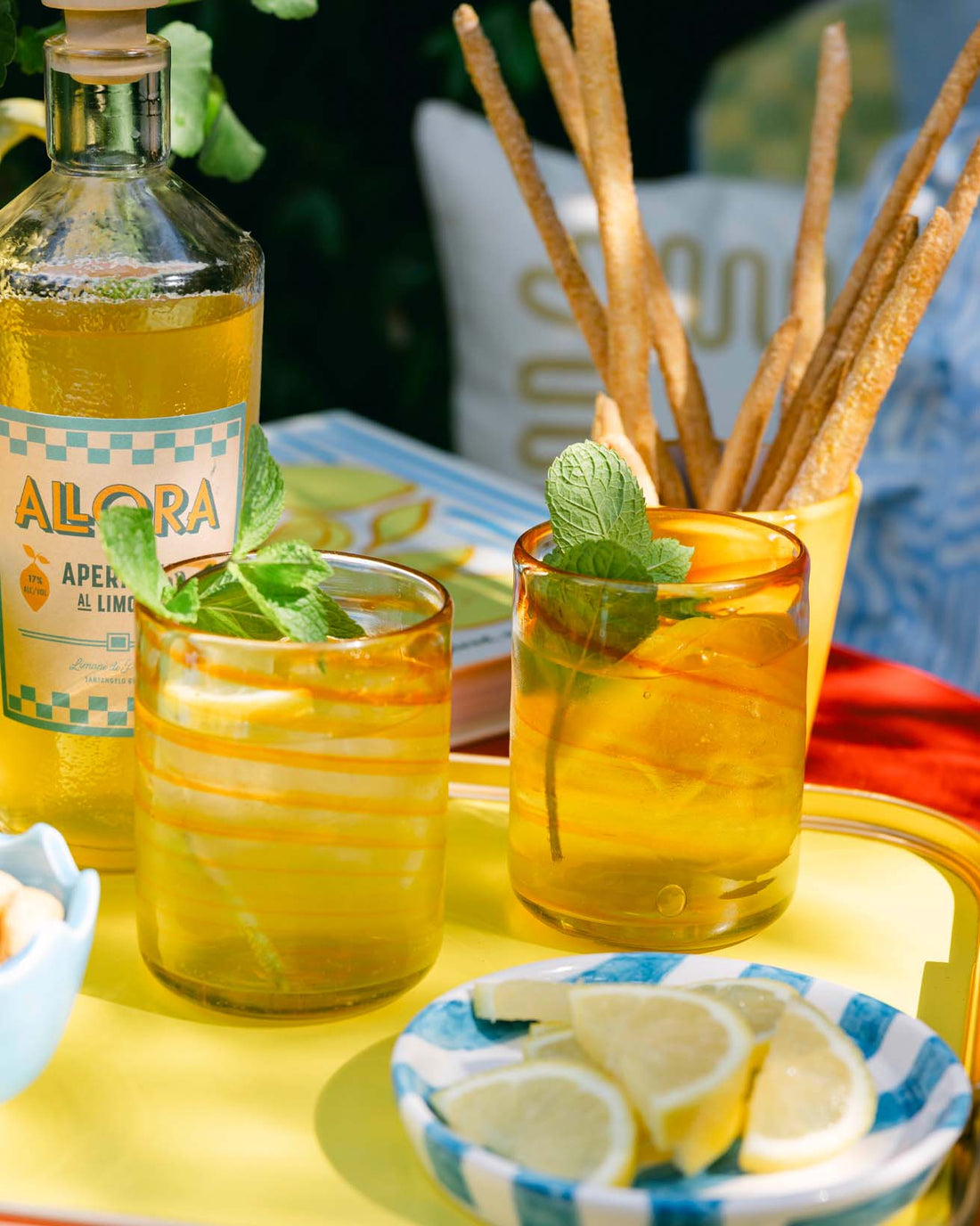 Two yellow glasses with mint leaves, a bottle labeled 'Allora', and lemon slices on a table. LAMP LDN LIVING