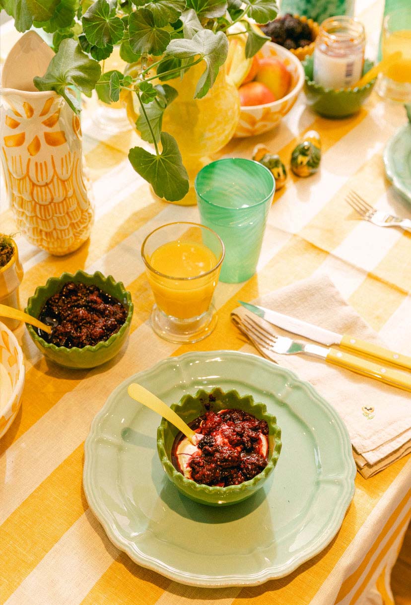 Dining table setup with green bowls, orange juice, and yellow utensils on a striped tablecloth. LAMP LDN LIVING