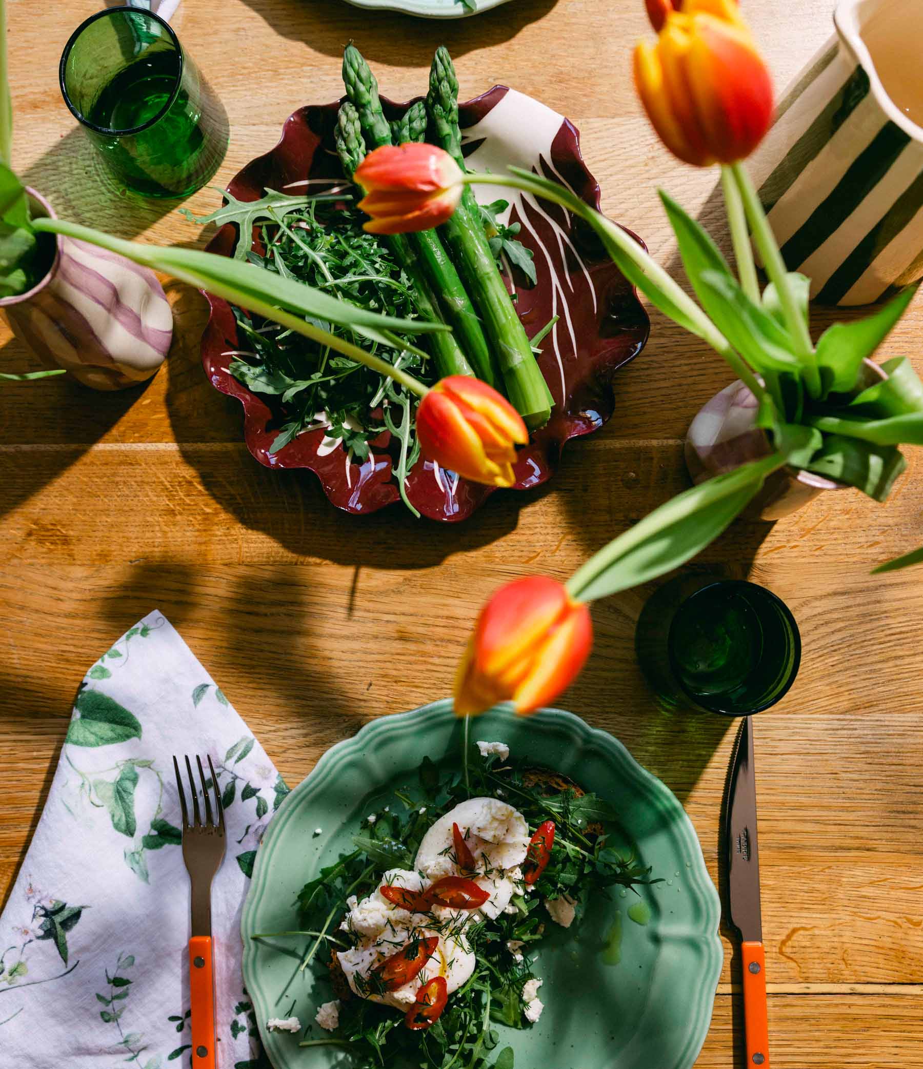 Dining table with plates of food, cutlery, tulips, and a striped vases on a wooden table. LAMP LDN LIVING